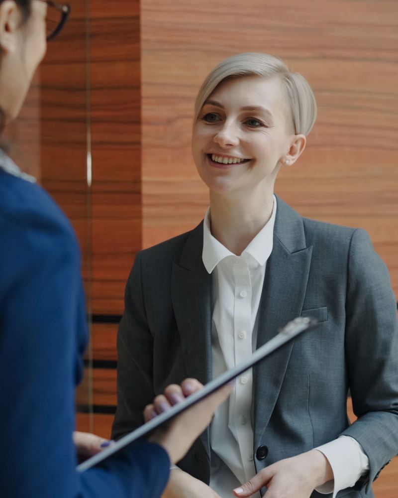 A smiling HR advisor in a gray blazer talking with a client who holds a clipboard, in an office with wood paneling